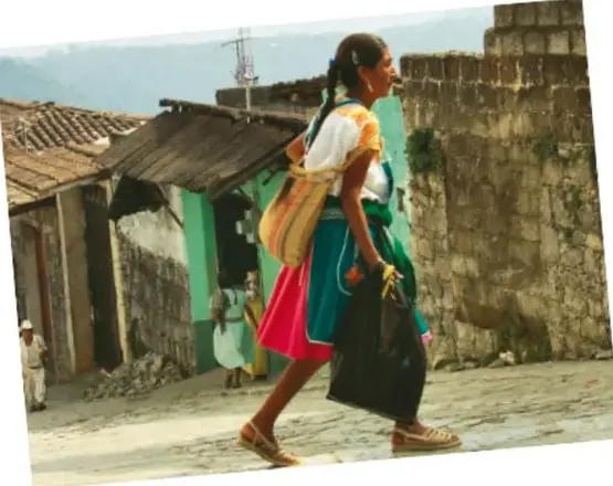 Una mujer caminando por una calle empedrada con un rebozo y una bolsa, junto a casas de adobe y una pared de piedra.