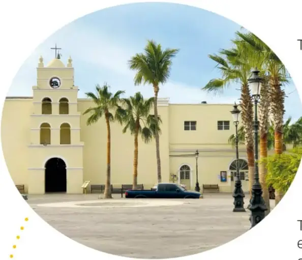 Fotografía del templo de la misión: edificio colonial claro con campanario, arcos y reloj, rodeado de palmeras y faroles, con una camioneta estacionada en la plaza.