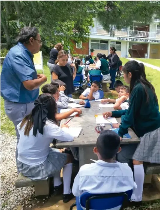 Fotografía de estudiantes de telesecundaria trabajando en grupos alrededor de mesas en el patio de la escuela, con un docente acompañándolos; en el fondo se ve la fachada con el rótulo "ESCUELA TELESECUNDARIA".
