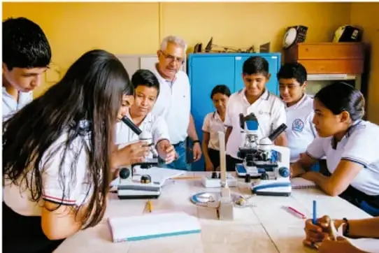Fotografía de estudiantes de telesecundaria y su docente trabajando con microscopios y material de laboratorio en una mesa de trabajo.