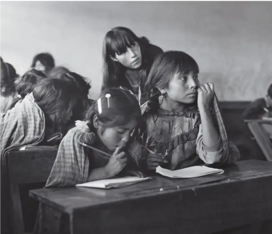 Fotografía en blanco y negro de niñas mazahuas en un salón de clases escribiendo en cuadernos sobre pupitres de madera, con una mujer joven detrás; Escuela mazahua, 1979, Estado de México, de Mariana Yampolsky (1925-2002).