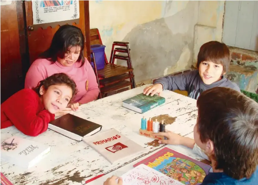 Fotografía de cuatro estudiantes sentados alrededor de una mesa de madera con libros, cuadernos y colores; uno de ellos sostiene un libro. Al fondo hay sillas apiladas y un cartel en la pared. Escenario escolar de trabajo en equipo.