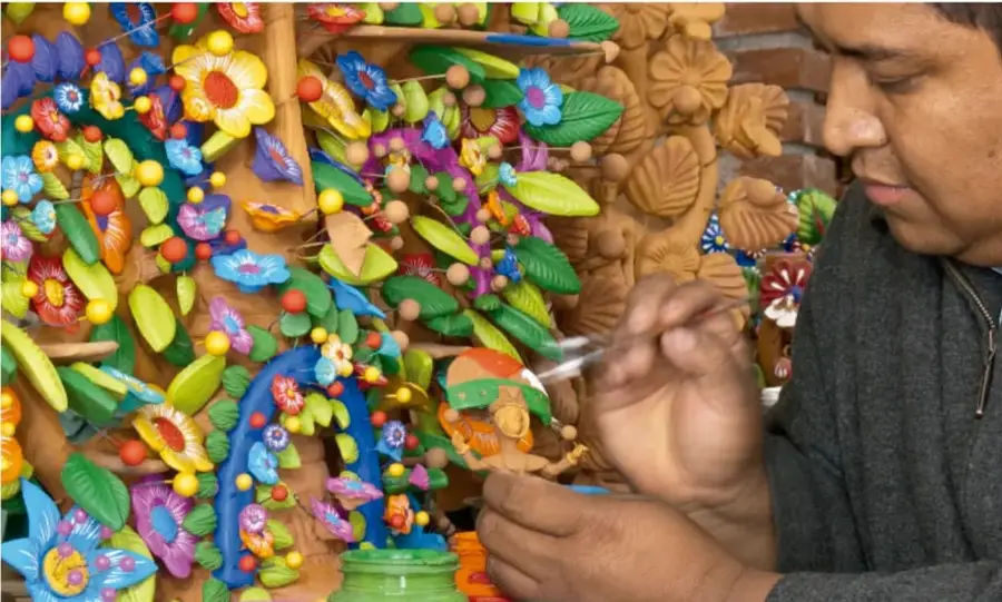 Artesano pintando con pincel piezas de barro; alrededor hay numerosas figuras de flores, mariposas y hojas coloridas. Crédito: Carlos Rahrin, LIDAM de México.