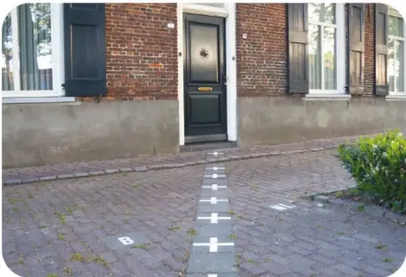 Banqueta de ladrillo frente a una casa de ladrillo con puerta negra y ventanas con contraventanas; una línea de cruces blancas pintadas en el piso marca la frontera entre Bélgica y los Países Bajos.