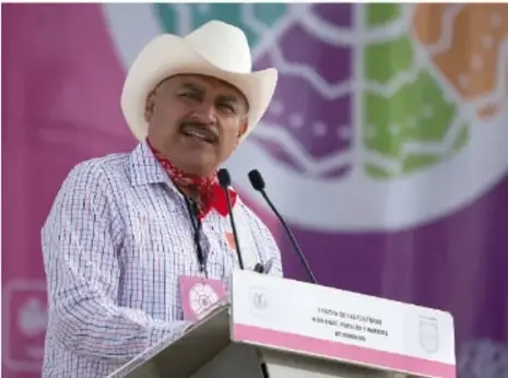 Fotografía de un hombre con sombrero blanco y pañuelo rojo al frente de un atril, en un evento con mantel rosa y logotipos institucionales.