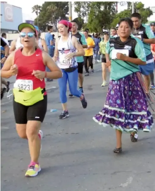 Fotografía de corredores participando en una carrera urbana: mujeres con ropa deportiva, chamarra tradicional y número de competencia, en una calle con árboles.