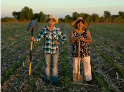 Fotografía de dos personas (hombre y mujer) con sombrero y herramientas agrícolas (pala y azadón) de pie en un campo agrícola con cultivo joven, iluminados por la luz del atardecer.