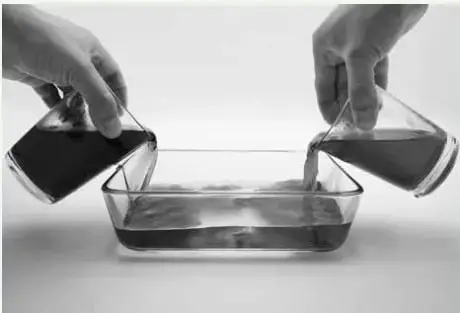 Dos manos vierten simultáneamente el contenido de dos vasos con líquido oscuro (teñido) por los extremos opuestos de un recipiente rectangular con agua.