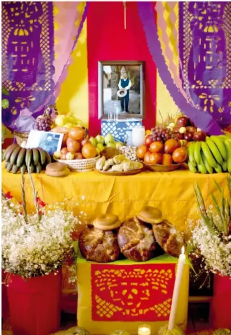 Ofrenda de Día de Muertos con mantel amarillo, frutas, pan de muerto, papel picado morado y amarillo, fotografía del difunto y velas.