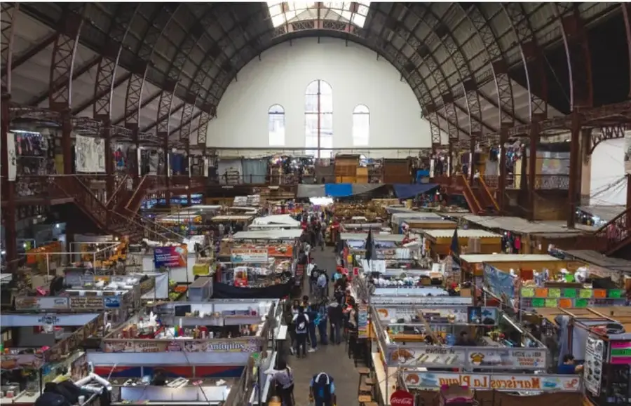 Fotografía del interior de un mercado público con estructura metálica y vitrales en el techo, pasillos con puestos de venta de frutas, verduras y abarrotes, y personas caminando entre los locales.