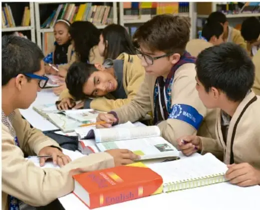 Fotografía de estudiantes de secundaria trabajando en grupo alrededor de una mesa con libros y cuadernos en la biblioteca escolar.