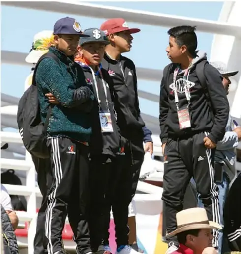 Grupo de estudiantes con chamarra deportiva y gafetes conversando en un evento deportivo al aire libre.