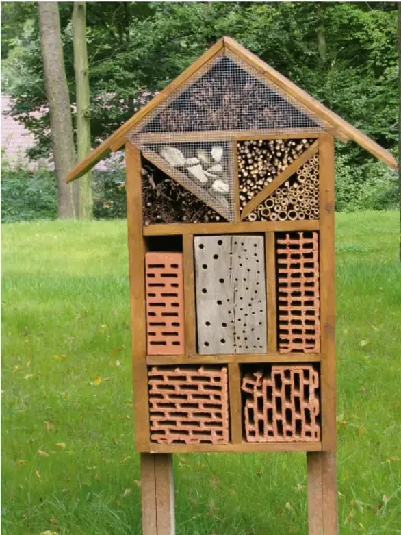 Fotografía de un hotel entomológico de madera con techo triangular a dos aguas, montado sobre dos postes. Contiene compartimentos con troncos perforados, ladrillos con huecos, bloques de madera con agujeros y tubos. Está en un jardín verde con árboles al fondo.