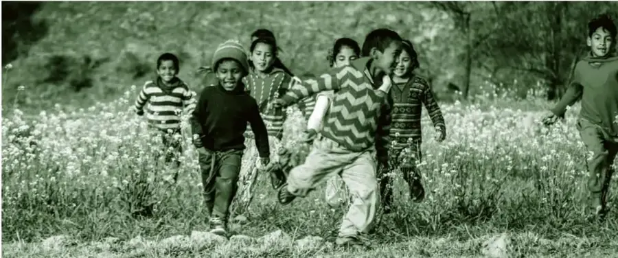 Fotografía en tonos verdes de varios niñas y niños corriendo por un campo de flores silvestres, algunos con gorros y suéteres de rayas, en plena persecución de juego.