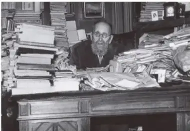 Fotografía en blanco y negro de un historiador de barba sentado en su escritorio, rodeado de montañas de libros y documentos.