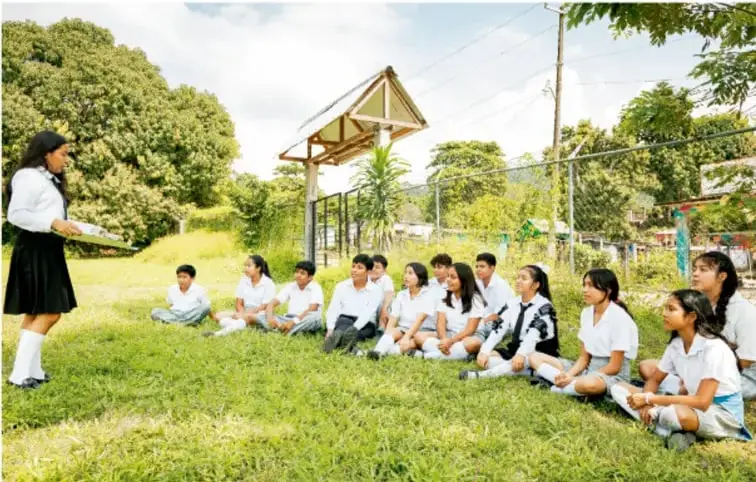 Fotografía de un grupo de estudiantes de telesecundaria con uniforme, sentados en el pasto al aire libre, atentos a una compañera de pie que sostiene un cuaderno y realiza la presentación del proyecto.