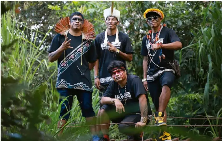 Fotografía de cuatro jóvenes indígenas en un entorno de vegetación alta; tres están de pie y uno arrodillado al frente. Visten camisetas negras con estampados, collares, pulseras y accesorios tradicionales; uno lleva un penacho o tocado con plumas y otro lentes de sol con un gorro decorado, combinando la indumentaria tradicional con ropa urbana.