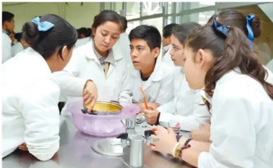 Fotografía de un grupo de estudiantes con bata blanca alrededor de una mesa de laboratorio; observan y manipulan material, uno de ellos sostiene un recipiente morado. Ilustra el trabajo científico colaborativo y la observación.