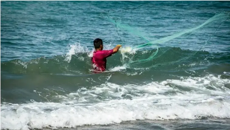 Fotografía de un pescador con camisa roja dentro del mar lanzando una red de pesca entre las olas.