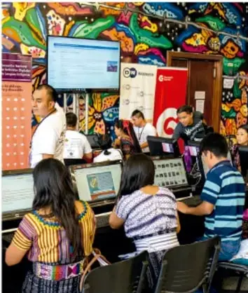 Fotografía de un salón con estudiantes y docentes frente a computadoras; al fondo un mural colorido de estilo mexicano con motivos populares.