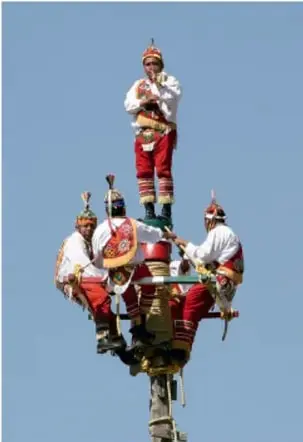 Fotografía de la Danza de los Voladores: cuatro hombres con trajes rojos y blancos y penachos, descendiendo desde un poste alto, como ejemplo de tradición y símbolo cultural de México.