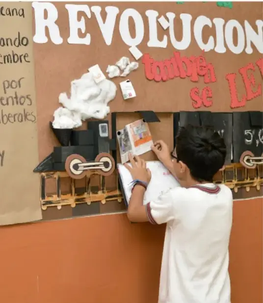 Niño trabajando en un periódico mural con la palabra 'REVOLUCIÓN' y figuras de trenes.