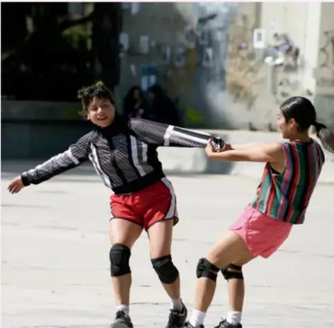 Dos niñas practicando un juego de tirón o arrastre con protecciones en rodillas, en un patio urbano.
