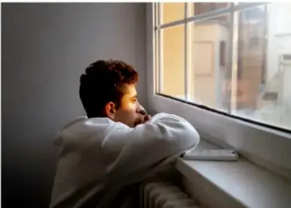 Fotografía de una persona joven reflexionando junto a una ventana, apoyada en el alféizar.