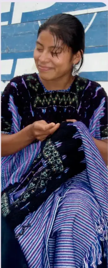 Fotografía de una mujer indígena joven, sonriente, sentada y sosteniendo un tejido textil de colores azul, morado y negro. Viste un huipil tradicional con bordados. Al fondo se ve una pared con pintura azul y blanca.