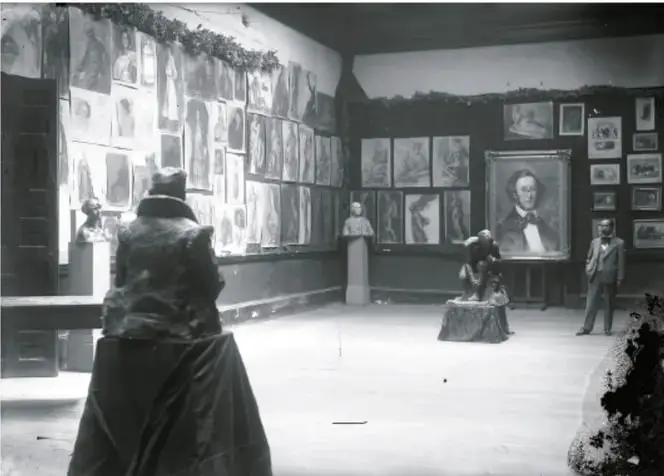 Fotografía en blanco y negro de una sala de la Academia de San Carlos. Se ven cuadros en las paredes, esculturas y una figura de espaldas en primer plano. Al fondo, una mujer de pie frente a un cuadro.