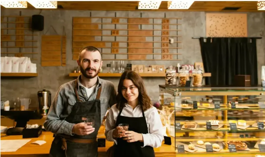 Fotografía de un hombre y una mujer con delantales detrás de un mostrador de cafetería o panadería, representando un pequeño emprendimiento.