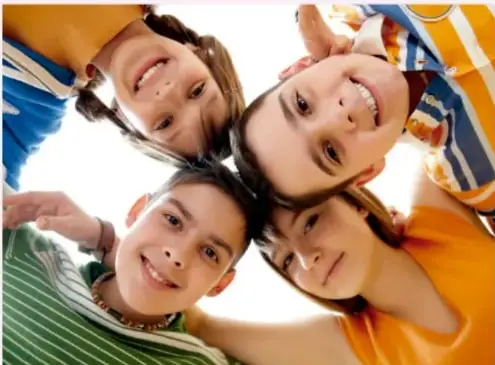 Fotografía de cuatro niños sonrientes vistos desde abajo (ángulo cenital), con la cabeza juntas formando un círculo, sobre fondo blanco; ilustra el tema de población infantil.