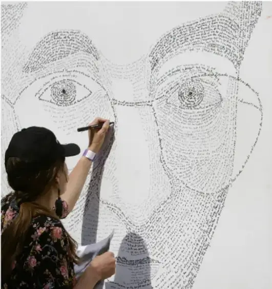 Fotografía de una persona que dibuja con texto un rostro gigante en una pared: un caligrama en proceso, donde las palabras forman los ojos y rasgos del rostro.