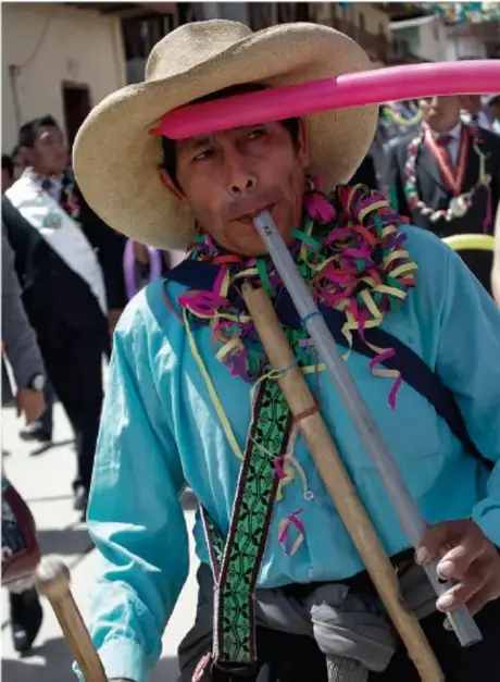 Fotografía de una persona con sombrero de ala ancha, ropa tradicional colorida y un instrumento de viento, en una calle con otras personas al fondo, representando una práctica cultural comunitaria.
