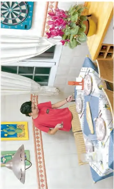 Fotografía de un comedor: un niño con polera roja junto a una mesa cubierta con mantel azul; sobre ella hay platos, un jarro y un tenedor. Se ven además una diana, un cuadro y una maceta con flores.