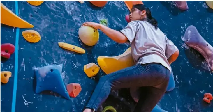Fotografía de una joven escalando un muro de escalada con presas de colores naranja, azul, rosa y amarillo