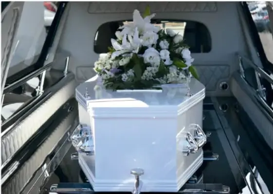 Fotografía de un féretro blanco con un arreglo floral de lirios dentro de una carroza fúnebre, símbolo de la mortalidad.