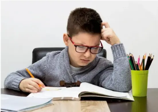 Fotografía de un estudiante con lentes escribiendo en un cuaderno, con lápices de colores en un vaso verde, mientras reflexiona sobre su trabajo escolar.