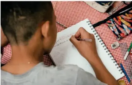 Niño escribiendo en un cuaderno sobre una mesa con mantel a cuadros.
