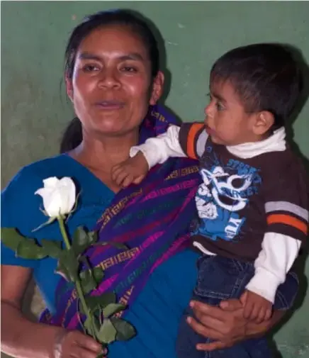 Fotografía de una mujer indígena con un niño en brazos sosteniendo una rosa blanca, en un contexto de conmemoración.