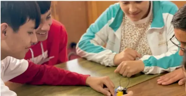 Fotografía de cuatro estudiantes alrededor de una mesa trabajando con un objeto pequeño, probablemente parte de su proyecto, con atención y colaboración.