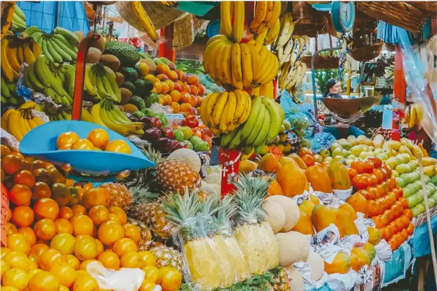 Puesto de mercado o tianguis con gran variedad de frutas: plátanos, naranjas, piñas, manzanas, jitomates y otras verduras, colgando y acomodadas en canastas.