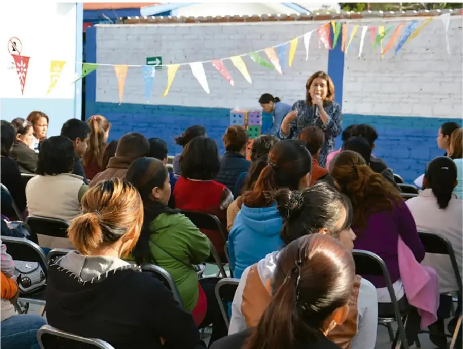 Fotografía de una reunión escolar: una mujer con micrófono habla a un público sentado en sillas, con banderines de colores colgados.