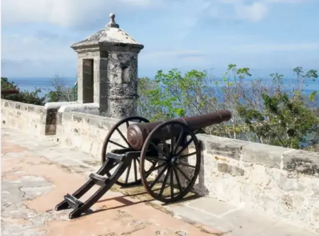 Cañón antiguo sobre cureña de madera en una muralla de piedra frente al mar, junto a una garita o cuerpo de vigía con arco.