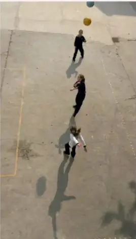 Vista aérea de niños jugando en una cancha con balones de colores, sombras largas en el piso.