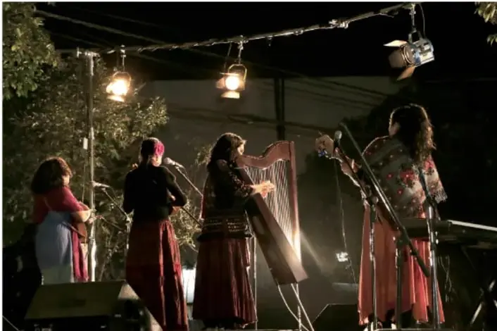 Fotografía nocturna de un evento comunitario: varias mujeres sobre un escenario iluminado con lámparas, una de ellas toca un arpa, en un ambiente de celebración tradicional.