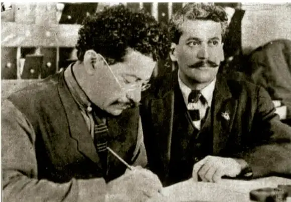 Fotografía en blanco y negro de dos hombres de la época revolucionaria. El de la izquierda, con anteojos y bigote, escribe sobre un escritorio; el de la derecha, también con bigote, observa. Podrían ser Francisco I. Madero y un colaborador.