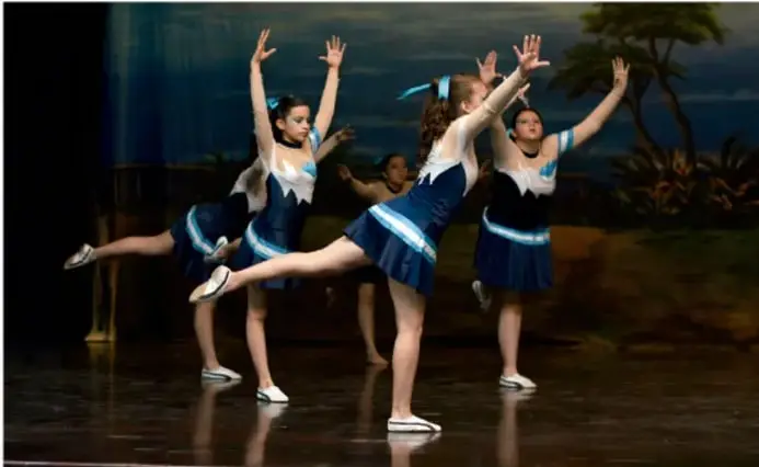 Fotografía de tres bailarinas con vestuario azul y blanco realizando una coreografía sobre un escenario con fondo de paisaje selvático.
