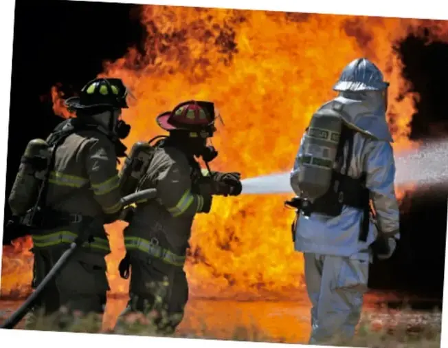 Fotografía de tres bomberos con equipo de protección combatiendo un incendio; uno de ellos sostiene una manguera mientras los otros observan.