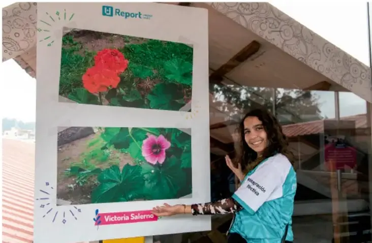Fotografía de una estudiante sonriente que presenta un cartel con dos fotografías de flores (una roja y una rosa), con un logotipo tipo 'Report' y una etiqueta con el nombre 'Victoria Salerno'.